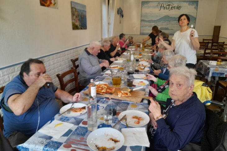 Anziani a tavola in un locale, mangiano pizza e bevono insieme